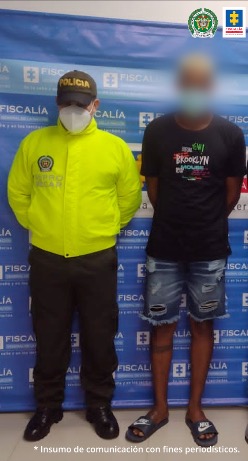 En la imagen se observa a un hombre de camiseta negra, bermuda de jean y chanclas negras, acompañado por un uniformado de la Policía Nacional, delante de un pendón de la Fiscalía General de la Nación.
