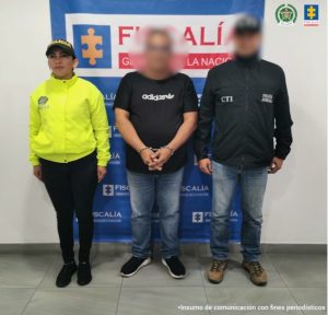 Se observa un hombre, capturado, en el centro, de frente. Sobre su rostro un efecto para difuminarlo. A un costado un funcionario del Cuerpo Técnico de Investigación CTI y al otro uno de Policía Nacional. Atrás un pendón de Policía Nacional.