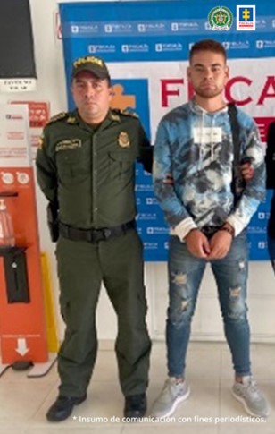 En la imagen se observa al capturado junto a un uniformado de la Policía Nacional. Detrás se observa el banner que identifica a la Fiscalía General de la Nación y la bandera de Colombia.