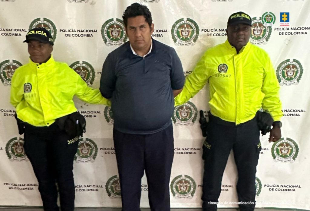 En la fotografía aparece de pie y con los brazos atrás Albert Giovanny Avilés Celis. Viste camisa de color azul oscuro, con cuello y manga corta; así como pantalón y zapatos negros. A sus extremos hay uniformados de la Policía Nacional y detrás de ellos hay un pendón institucional de la Policía.