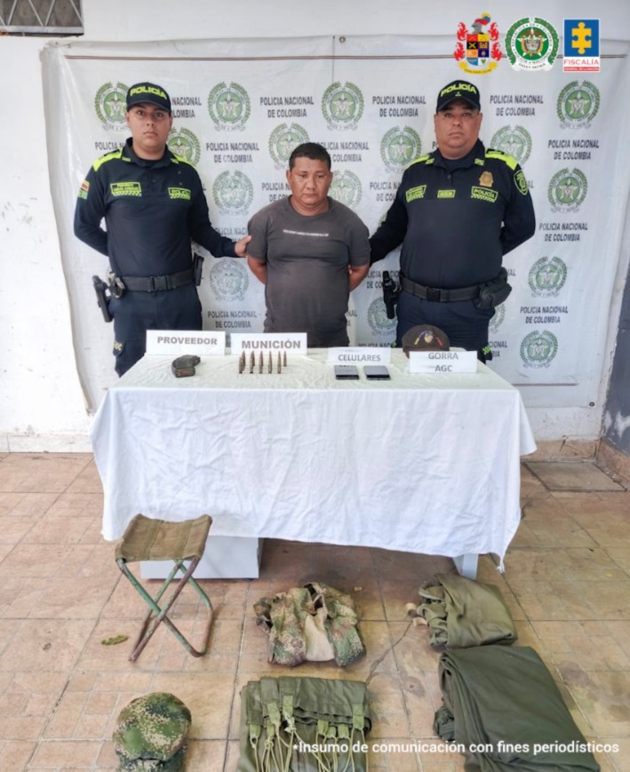 En la imagen aparece una persona de camiseta gris, entre dos uniformados de la Policía y frente a una mesa con munición, celulares y otros objetos.