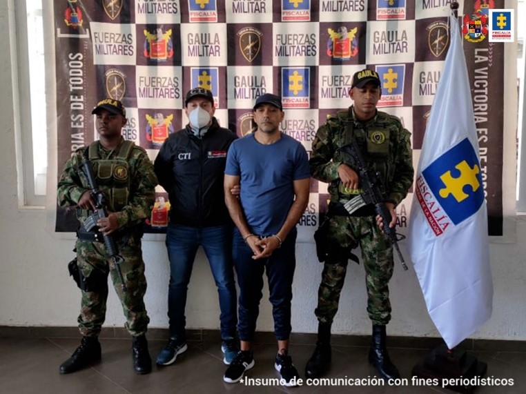 En la fotografía se observa el capturado junto a un investigador del CTI y dos uniformados del Gaula Militar. Se observa la bandera de la Fiscalía y en la parte posterior se encuentra un banner con los logos del Gaula Militar y de la Fiscalía General de la Nación.