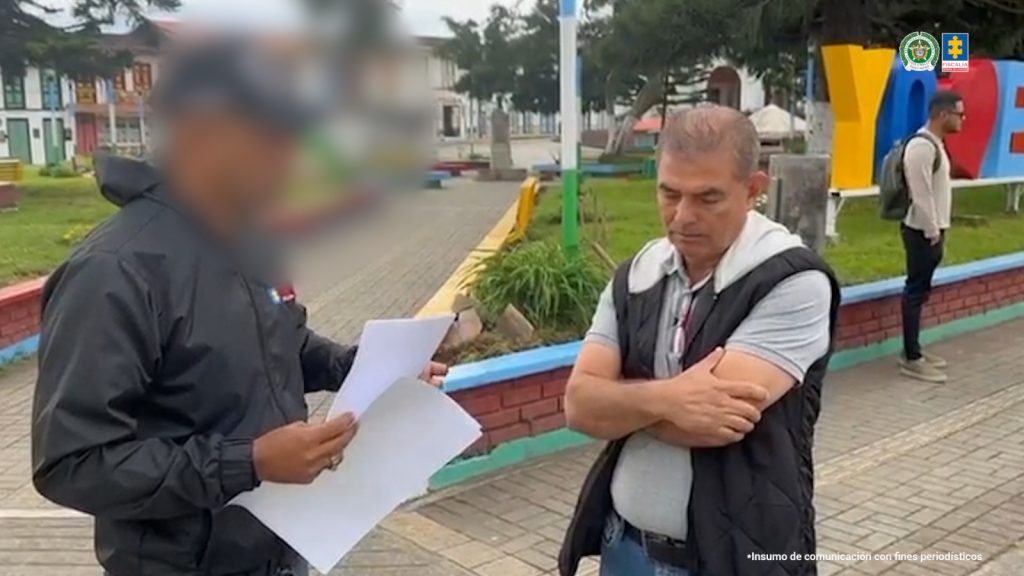 La fotografía está tomada en exteriores. Se aprecia de pie con los brazos cruzados a Olaín Salazar Ramírez. Viste con camisa gris, de manga corta; chaleco de color negro y jean azul con correa negra. Frente a él hay un servidor del CTI de la Fiscalía en acción de lectura de unos documentos que tiene en sus manos.