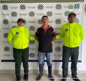 Se observa al capturado de frente y a cada costado un funcionario de Policía nacional. Atrás, un pendón de Policía Nacional.