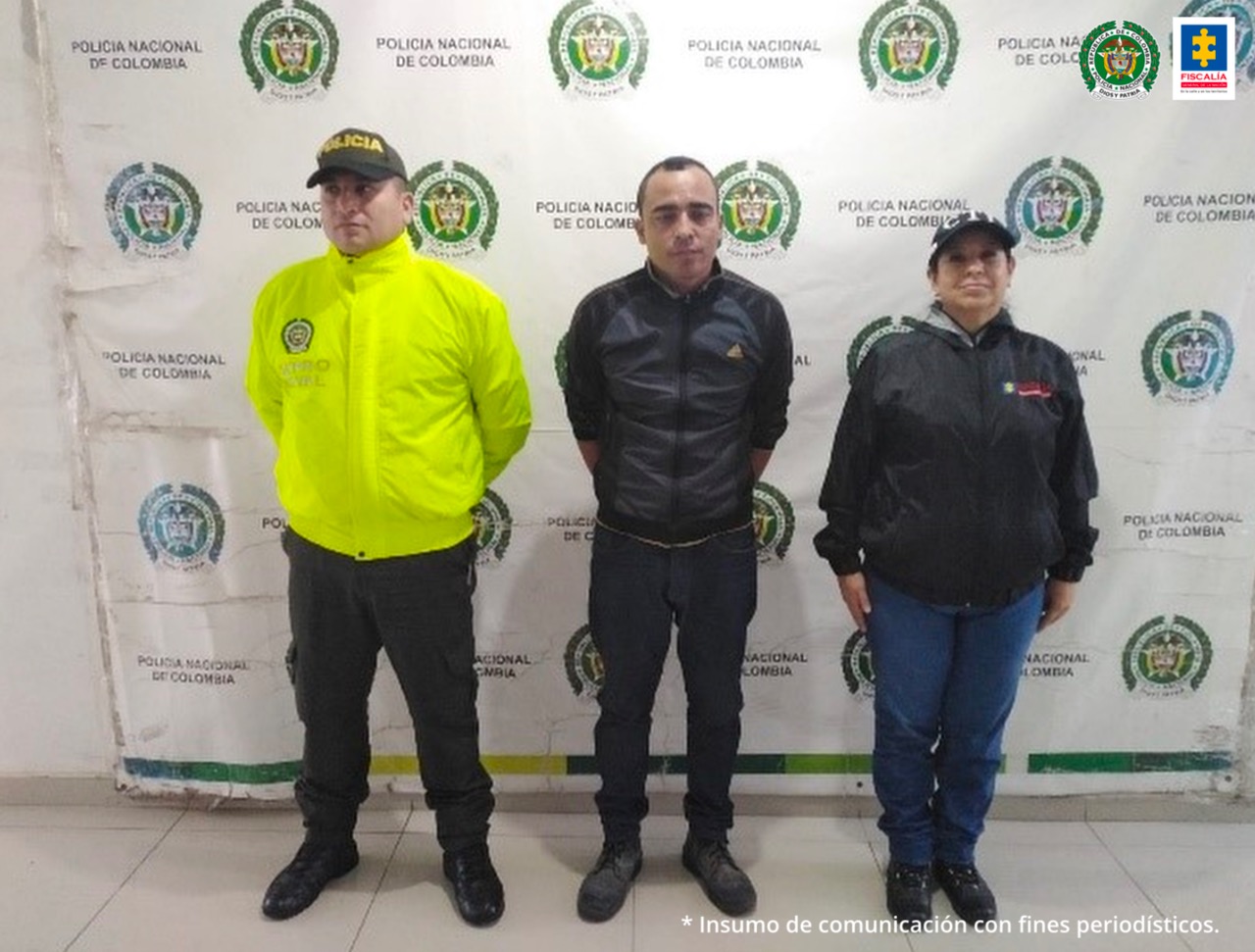 En la foto aparece Alejandro Antonio Upegui Cortez, de cuerpo completo y con los brazos atrás; vestido con chaqueta, pantalón y zapatos negros. Al lado izquierdo de él hay un uniformado de la Policía Nacional y a su izquierda una servidora del CTI de la Fiscalía. Detrás de ellos hay un pendón institucional de la Policía Nacional.