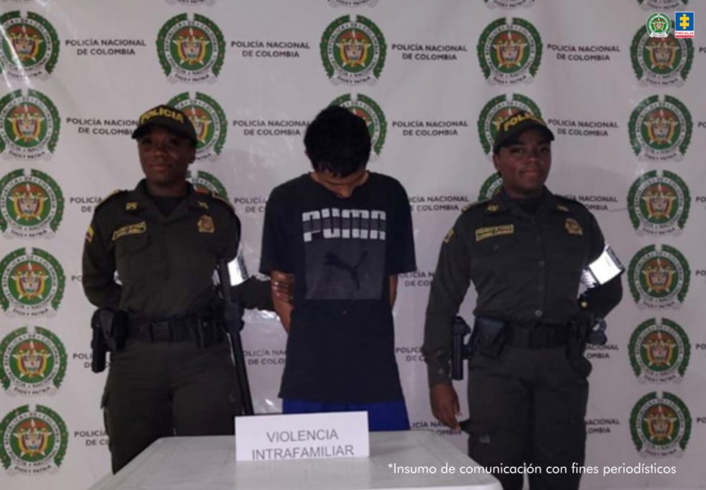 En la fotografía se aprecia de pie, con la cabeza agachada y los brazos atrás a Alejandro Morales Pérez. Viste con camiseta de color negro con estampado de la palabra ‘Puma’ a la altura del pecho; jean azul oscuro A sus extremos hay dos uniformadas de la Policía Nacional. Delante de ellos hay una superficie blanca y sobre ella hay un letrero que dice: ‘Violencia intrafamiliar’. Detrás de ellos hay un pendón institucional de la Policía.