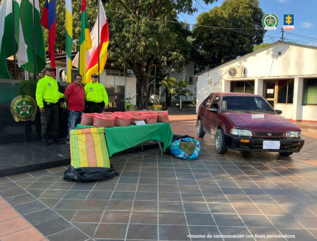 Se observa al capturado custodiado por dos uniformados de la Policía Nacional. En la imágen se puede apreciar una mesa con el estupefaciente incautado y el vehículo.
