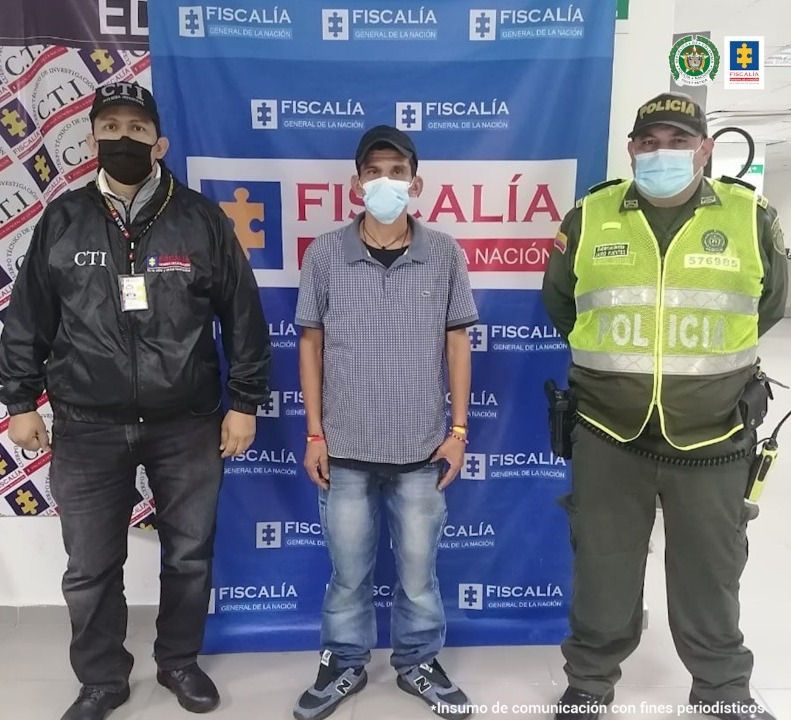 En esta foto se ve un hombre joven con tapaboca, camiseta, tenis y jean custodiado por investigador del CTI de la Fiscalía y un uniformado de la Policía Nacional, ambos con tapaboca. Destras de ellos hay un banner de la Fiscalía.