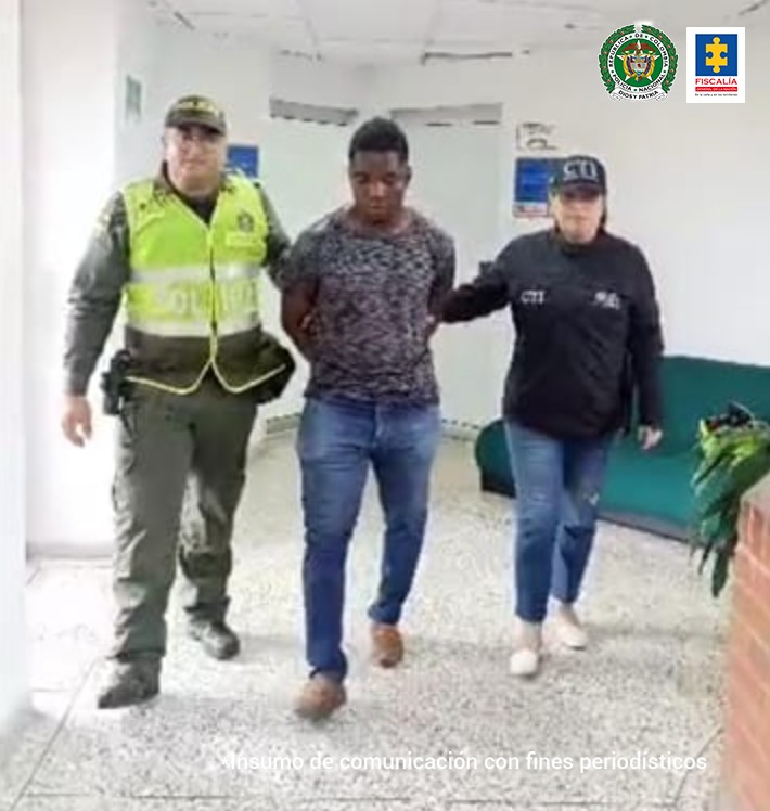 En la fotografía aparece en un pasillo Alexander Caicedo Camilo en acción de caminata, con los brazos atrás. Viste camiseta estampada, jean azul y zapatos color miel. Al lado derecho de él hay un uniformado de la Policía Nacional. Al lado izquierdo hay una servidora del CTI de la Fiscalía.