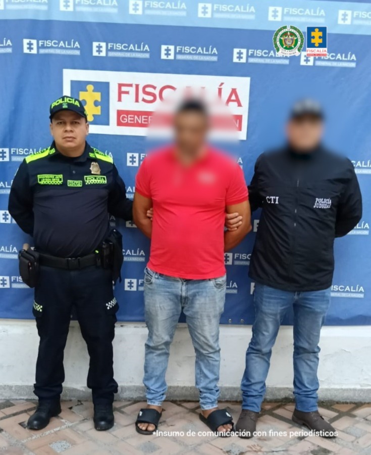 Se visualiza al capturado con el rostro difuminado junto a un investigador del CTI y un uniformado de la Policía Nacional. Detrás de ellos el banner que identifica a la Fiscalía General de la Nación