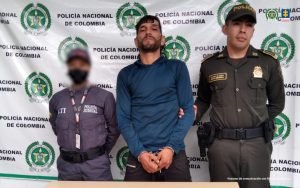 En la fotografía aparece de pie, con los brazos adelante y esposado Alexander Osorio López. Viste con buso azul oscuro y pantalón negro. Usa barba y bigote. En el extremo derecho de él hay una servidora del CTI de la Fiscalía y en el derecho un uniformado de la Policía Nacional. Delante de ellos hay una superficie y sobre ella hay un letrero que dice: ‘Orden judicial’. Detrás de ellos hay un pendón institucional de la Policía.