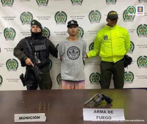 En la foto se aprecia a Alexis Hoyos Henao, alias Tuto, de pie y con los brazos atrás. Viste camiseta gris de manga corta con estampado circular negro a la altura del pecho. A lado y lado de él hay uniformados de la Policía Nacional. Delante de ellos hay una superficie marrón y sobre ella se aprecia munición y un arma de fuego.