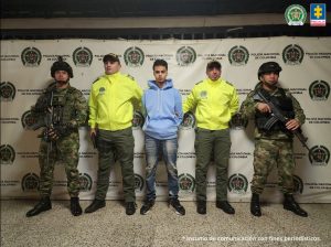 Se visualiza al capturado de frente, esposado con las manos atras junto a dos uniformados de la Policía Nacional y dos uniformados del ejercito nacional. Detrás de ellos el banner que identifica a la Policía Nacional