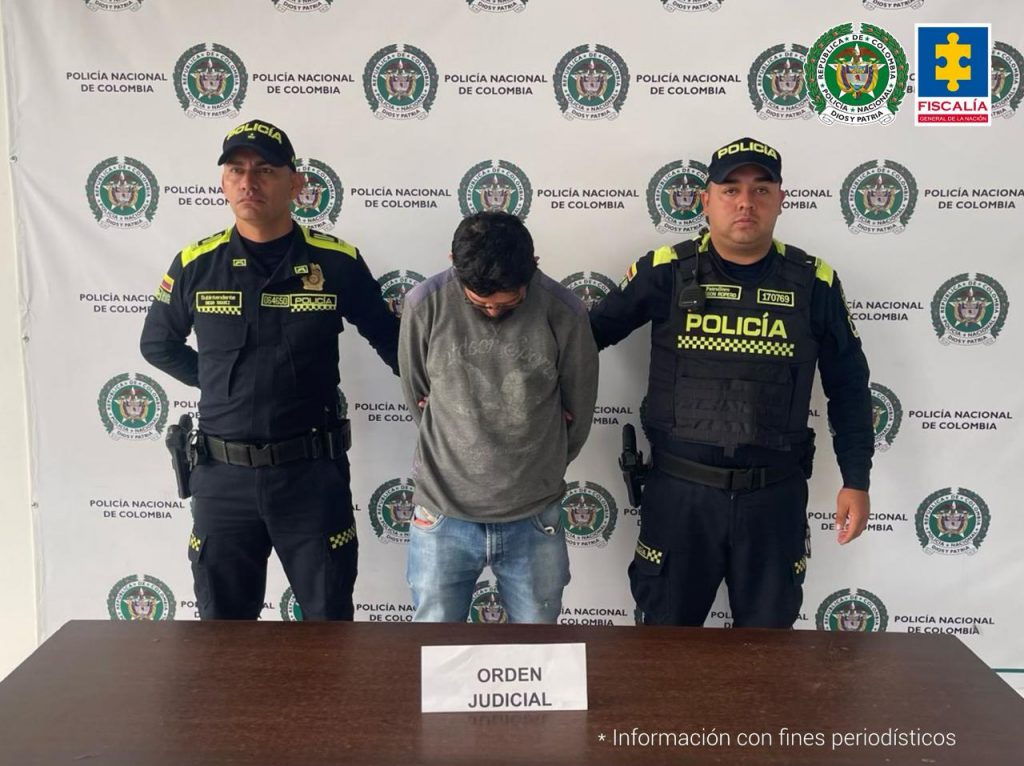 El capturado se encuentra de frente y custodiado por dos uniformados de la Policía Nacional. Detrás de la foto se observa un backing de la Policía Nacional.