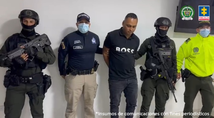 El capturado aparece en medio de la imagen, rodeado de personal de Goes, Policía e Interpol. Está esposado con las manos atrás.