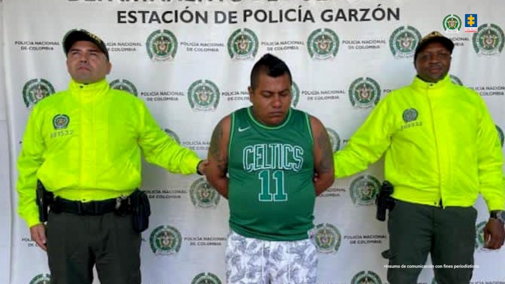 En la foto se observa al agresor quien tiene un buzo verde y unas bermudas blancas con estampados y se encuentra en custodia por dos uniformados de la Policía Nacional. Detrás hay un backing de la Policía Nacional.