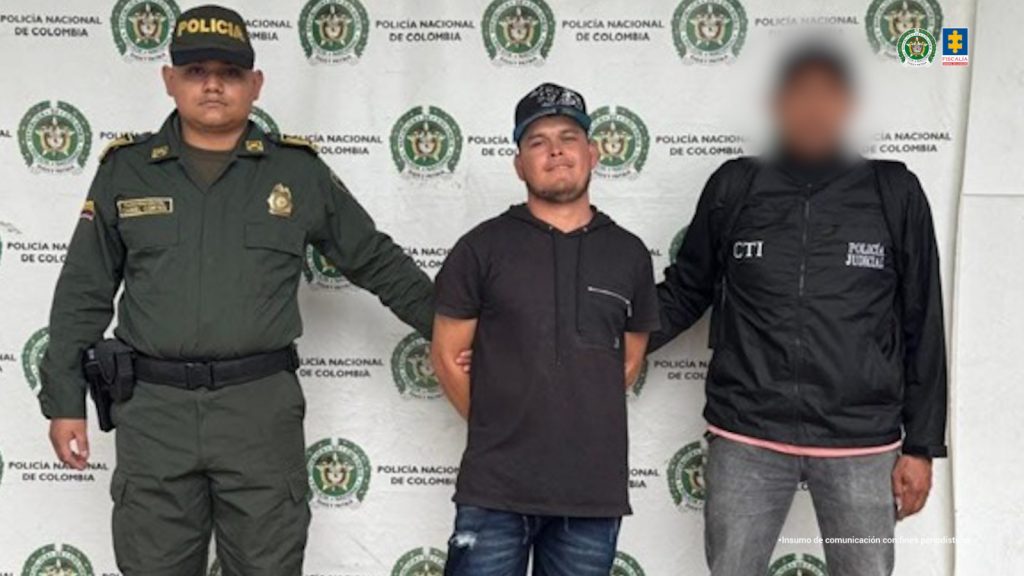 En la foto se observa al agresor quien tiene una gorra y se encuentra en custodia por un servidor del CTI y un uniformado de la Policía Nacional. Detrás hay un backing de la Policía Nacional.