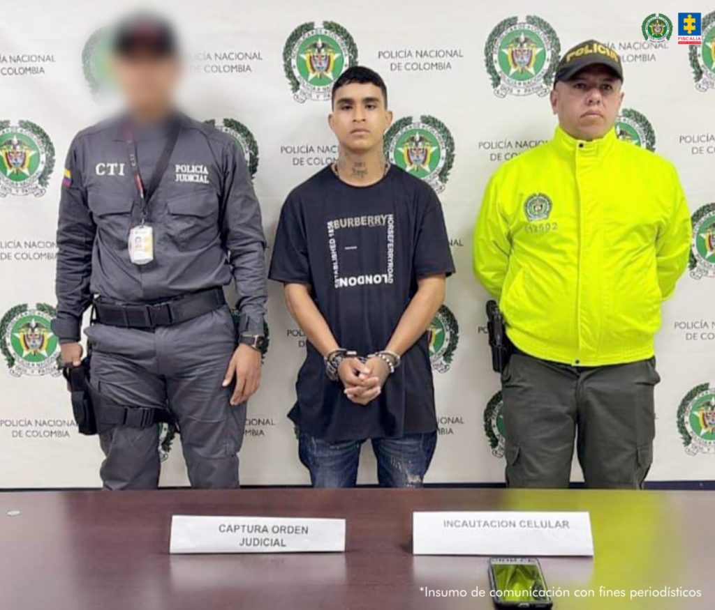 En la fotografía aparece el hombre junto a un servidor del CTI y un uniformado de la Policía Nacional. En la parte posterior está una mesa con un teléfono celular.