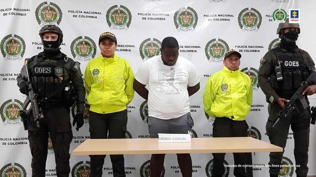 En la fotografía se observan cincos personas, una e ellas el capturado los cuatro restantes uniformados de la policía. En la parte posterior un backing con escudos de la Policía Nacional.