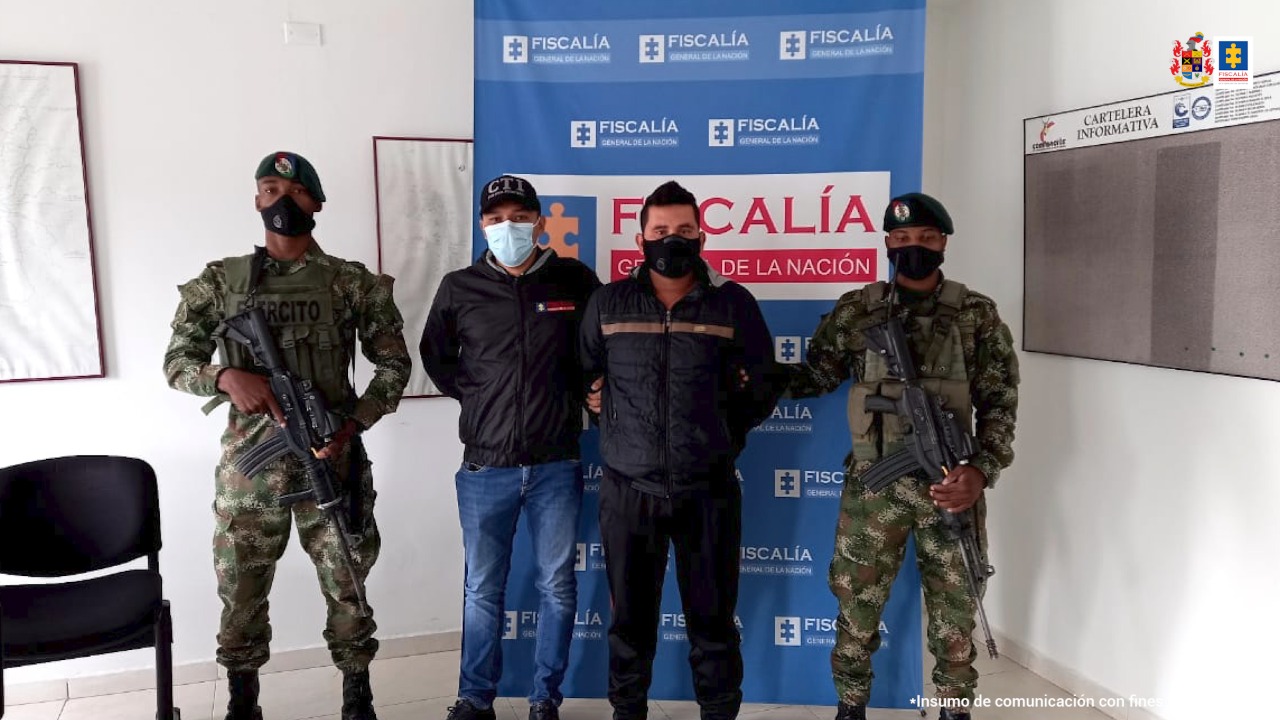 En la foto se observan cuatro personas, una de ellas el capturado, dos integrantes del Ejército Nacional y un integrante del Cuerpo Técnico de Investigación. En la parte posterior se encuentra un backing de la Fiscalía General de la Nación.