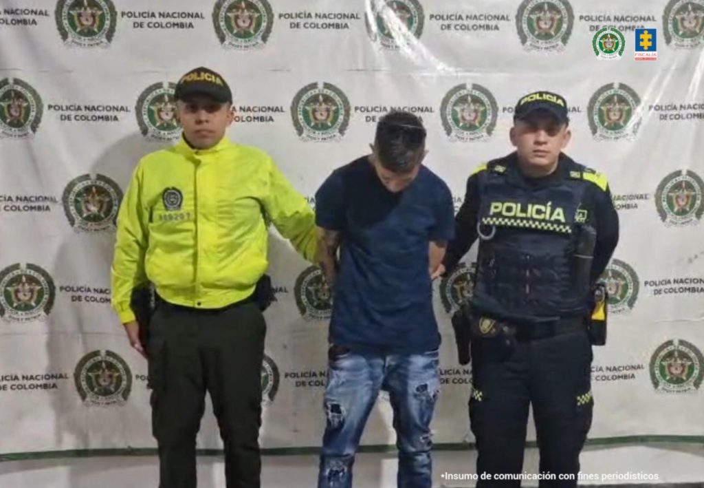 El procesado se encuentra en custodia por dos uniformados de la Policía Nacional. El capturado tiene un buzo color azul oscuro y un jean. Detrás se observa un backing de la Policía.