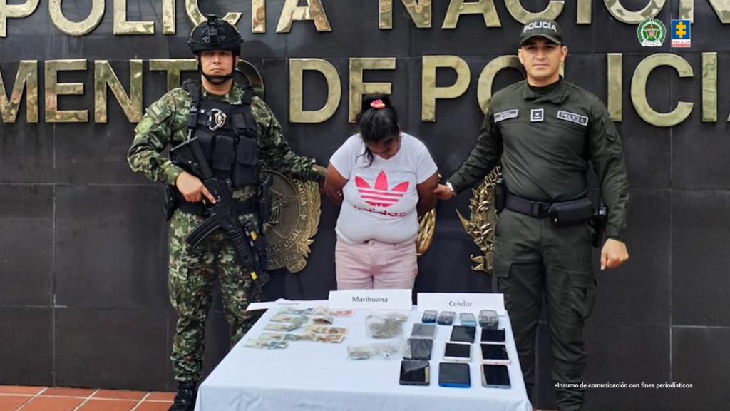 En la foto aparece la capturada custodiada por uniformados de la Policía Nacional.  En la foto también aparece lo que se le incautó durante la diligencia de allanamiento y registro.