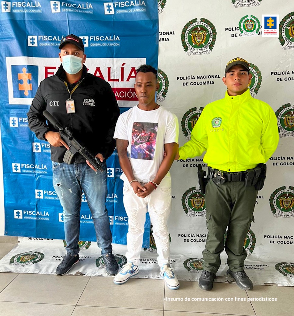 En la foto aparece alias ‘Morocho’, vestido con camiseta y pantalón blancos, con las manos adelante esposadas. Al lado derecho de alias ‘Morocho’ aparece un servidor del CTI y a su izquierda uno de la Policía Nacional. Detrás de ellos aparecen los pendones institucionales de Fiscalía y Policía.