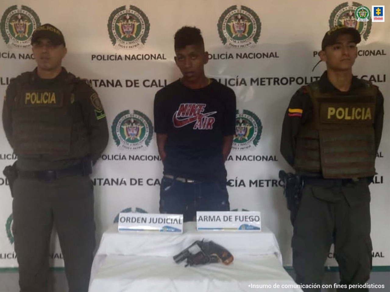 El capturado aparece con las manos esposadas en su espalda, frente a él una mesa con los objetos incautados y dos policías a cada costado.