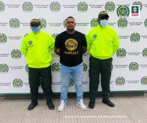  En la fotografía se observan tres personas una de ellas el capturado, junto a dos integrantes de la Policía Nacional, en la parte posterior se onbserva un backing con escudos de la Polcía Nacional. 