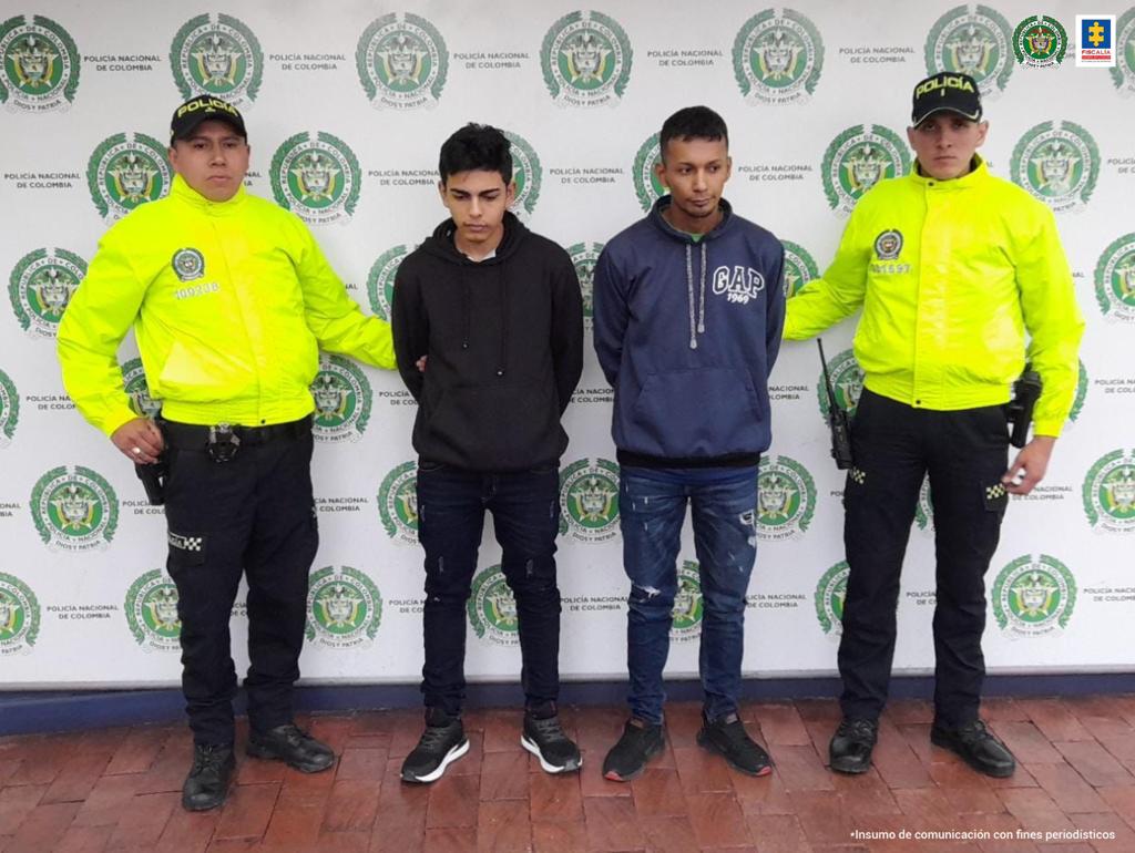 En la fotografía se observan cuatro personas, dos de ellas los capturados y dos funcionarios de la Policía Nacional. En la parte posterior se encuentra un backing con escudos de la Policía Nacional.