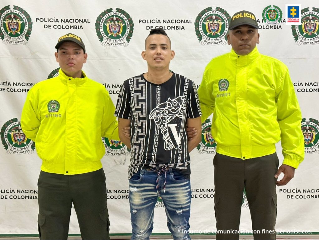 En la fotografía se observan tres personas, dos de ellas integrantes de la Polcía Nacional y uno de ellos el capturado. En la parte posterior se observa un backing con escudos de la Policía Nacional
