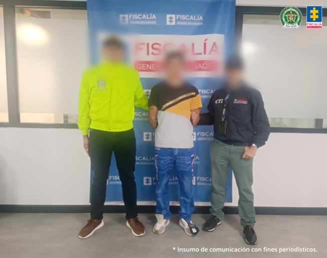 Se observa al capturado con el rostro difuminado junto a un uniformado de la Policía Nacional y un investigador del CTI. Detrás de ellos se ubica el banner institucional de la Fiscalía General de la Nación.