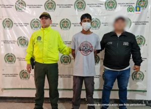 El capturado se encuentra de frente y custodiado por un servidor del Cuerpo Técnico de Investigación CTI, y un uniformado de la Policía Nacional. Detrás hay un backing de la Policía y Ejército. El capturado tiene una camiseta de color gris, un jean oscuro y tiene tapabocas