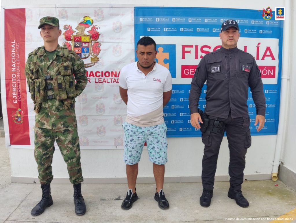 En la fotografía aparece un capturado junto a personal de Fiscalía General de La Nación y  Ejército  Nacional. En la parte posterior de la imagen se ven banners de Ejército Nacional y la Fiscalía General de la Nación.    
