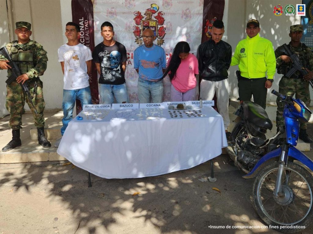 En la fotografía aparecen cuatro hombres y una mujer capturados, acompañados de dos uniformados de la Policía y el Ejército Nacional. En la parte posterior un banner con logos de la entidad.