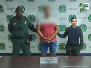 En la fotografía se aprecia a Ánderson Ochoa Bríñez. Viste camiseta de manga corta blanca en la parte superior y roja del pecho hacia abajo. Al lado y lado de él hay uniformados de la Policía Nacional. Delante de ellos hay una superficie color marrón y sobre ella hay un letrero que dice ‘Violencia intrafamiliar’. Detrás de ellos hay un pendón institucional de la Policía.
