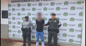 capturado de frente manos atrás esposado, custodiado por dos policías. Detrás banner de la Policía Nacional.