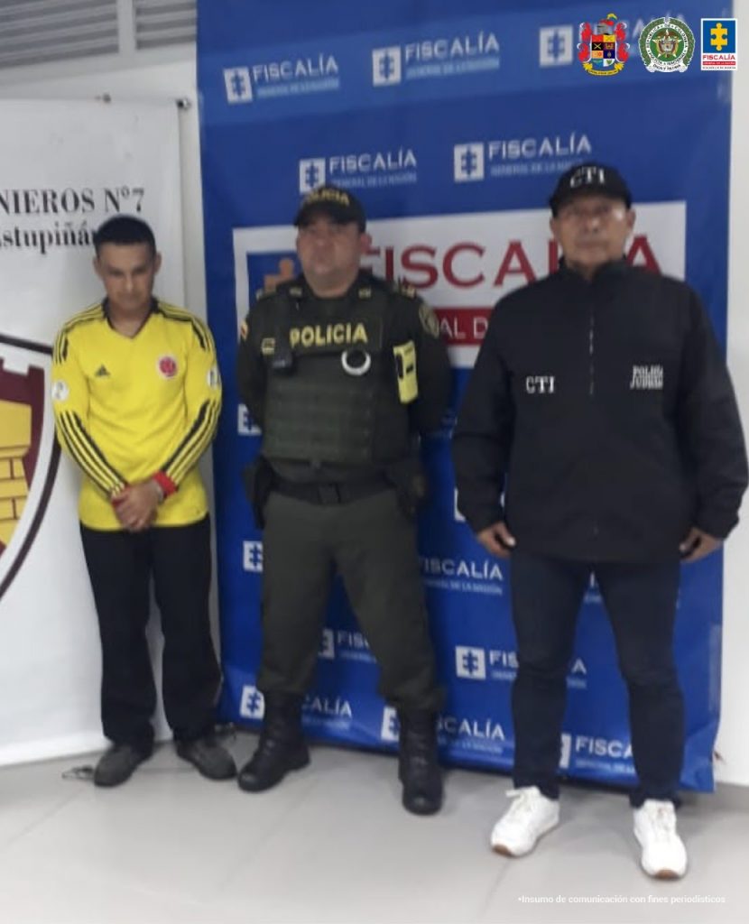 En la foto aparece a la izquierda el detenido de frente custodiado por un uniformado de la Policía y un investigador del CTI de la Fiscalía a su derecha detrás de ellos el banner de Fiscalía y parte del banner de Ejército