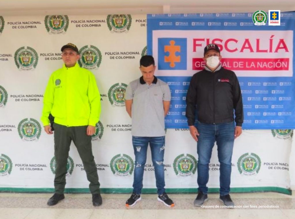 En la fotografía aparece un capturado junto a personal de Fiscalía General de La Nación y Policía Nacional. En la parte posterior de la imagen se ven banners de Policía Nacional y la Fiscalía General de la Nación.