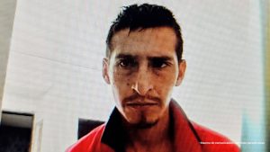 En la foto se ve a una persona de frente, con expresión seria y mirada directa a la cámara. Tiene el cabello oscuro, corto y peinado hacia atrás, y lleva un bigote fino con una pequeña perilla. Viste una prenda roja con cuello oscuro, que parece una chaqueta o camisa.