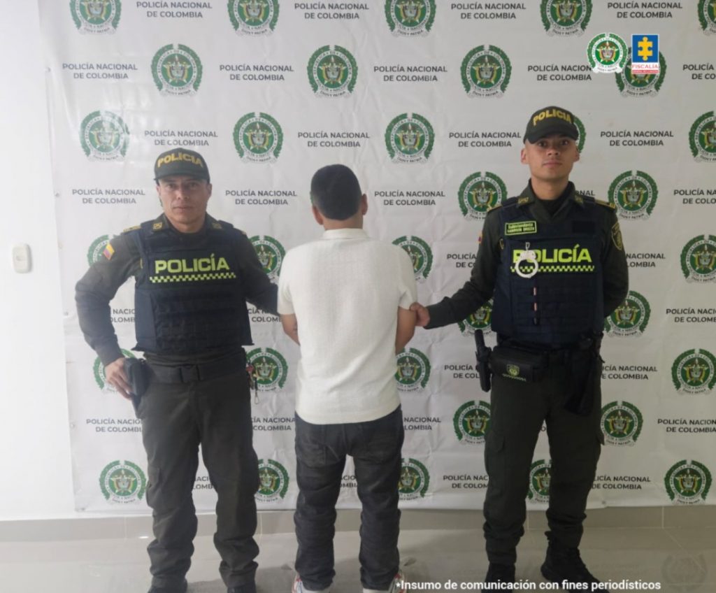Se observa al capturado de espaldas. Viste una camisa blanca y pantalón negro. A cada lado lo custodia un funcionario de la Policía Nacional. Al fondo, un pendón de Policía Nacional