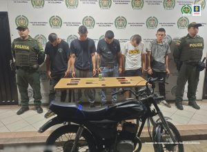 Se observan cinco detenidos en fila y a cada extremo un uniformado de Policía Nacional. Al fondo un pendón de Policía Nacional, al frente una mesa con el material incautado y adelante una motocicleta