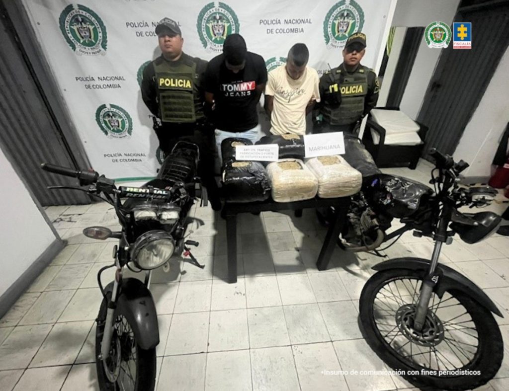 En la fotografía aparecen de pie, con los brazos atrás y la cabeza inclinada hacia adelante, Andrey Felipe Vásquez Saldarriaga y Luis Miguel Estrada Namundia. Uno, viste con camiseta de mangas cortas y de color negro, con estampado a la altura del pecho, así como jean azul claro. El otro imputado viste con camiseta de mangas cortas, de color beage y con estampado a la altura del pecho. En los extremos de ellos hay uniformados de la Policía Nacional. Delante de ellos hay una mesa y sobre ella hay paquetes envueltos en plástico negro y otros en papel con plástico transparente. Sobre ellos hay dos letreros (uno nítido y otro borroso) que dicen: ‘Marihuna’ y ‘Art. 376 Fabricación o porte de estupefacientes’. En los extremos también hay dos motocicletas. En la parte de atrás hay un pendón institucional de la Policía.