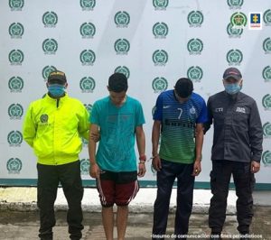 En la fotografía aparecen dos hombres capturados junto a un funcionario de la Policía Nacional y del CTI. En la parte posterior aparece un banner de la Policía Nacional.