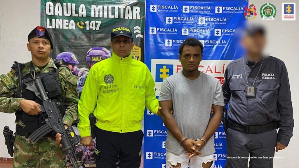 En la imagen aparece una persona de camiseta gris y bermuda blanca, entre tres uniformados del CTI, la Policía y el Ejército Nacional.