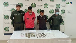 En la fotografía aparecen de pie y con los brazos atrás Anthonis Eloy Cortez Abache y Yeimi Manuel Freides Albornoz. Uno, viste con buso de color rojo y con estampado negro a la altura del pecho; así como pantalón oscuro. La otra persona viste con camisa de cuello blanco, mangas cortas de color negro y pantalón negro. En sus extremos hay uniformados de la Policía. Delante de ellos hay una superficie con mantel blanco y sobre ella se aprecian gran cantidad de bolsas pequeñas con contenido claro, rosado, vegetal y café. Detrás de ellos hay un pendón institucional de la Policía.