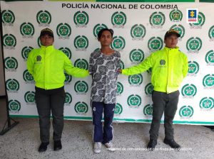  El detenido esposado custodiado por dos uniformados de la policía y detrás banner de Policía.