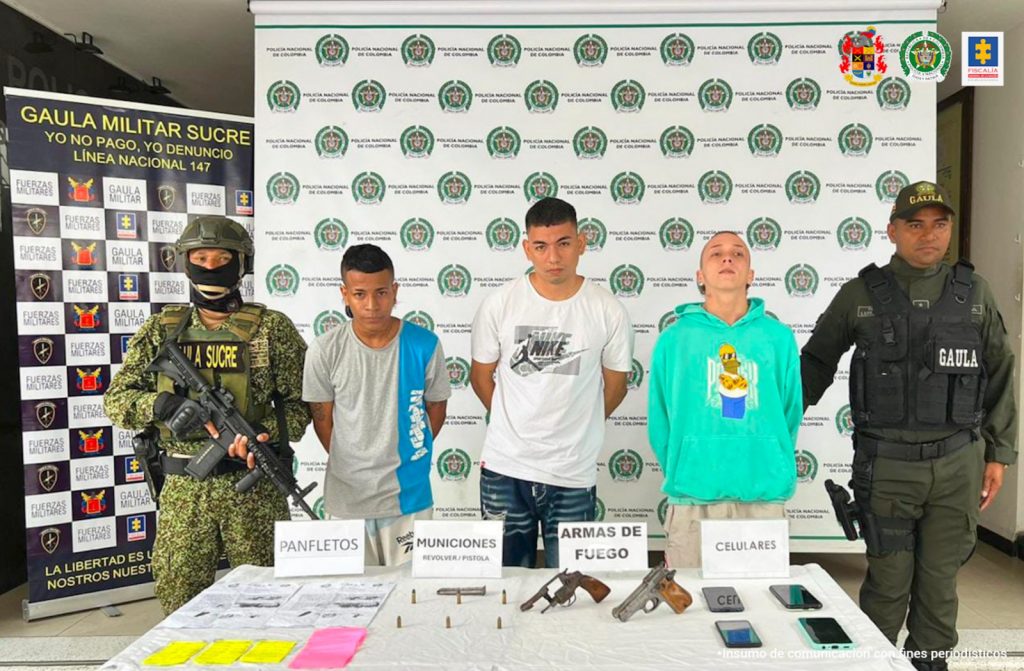 En la imagen aparecen tres personas, entre dos uniformados y frente a una mesa con armas de fuego y documentos.