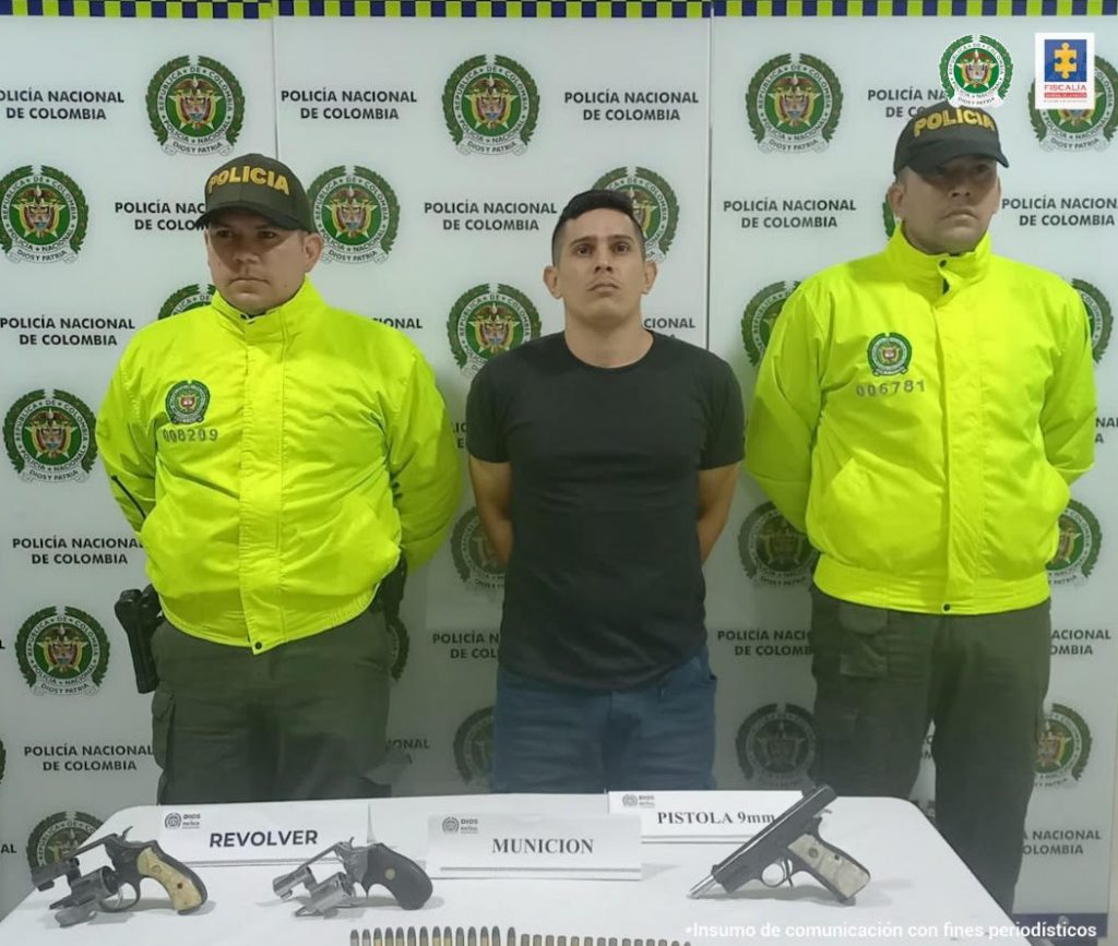 En la foto se aprecia al hoy asegurado siendo custodiado por un miembro de la Policía. Al frente, sobre una mesa hay tres armas de fuego de corto alcance.