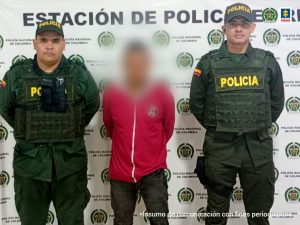 capturado de frente manos atrás esposado, custodiado por dos uniformados de la Policía. Detrás banner de la Policía Nacional.